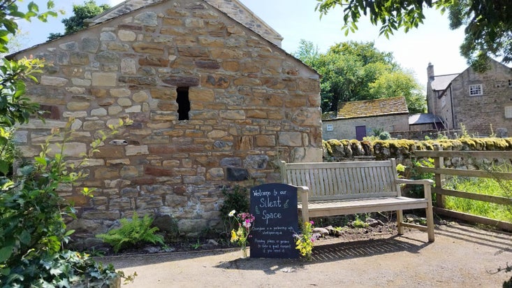 A wooden bench sits in front of an old stone building surrounded by greenery. A blackboard next to it says welcome to our silent space